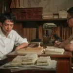 Two men sit at a wooden desk cluttered with stacks of papers in a dim office or archive, each examining documents.