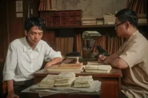 Two men sit at a wooden desk cluttered with stacks of papers in a dim office or archive, each examining documents.