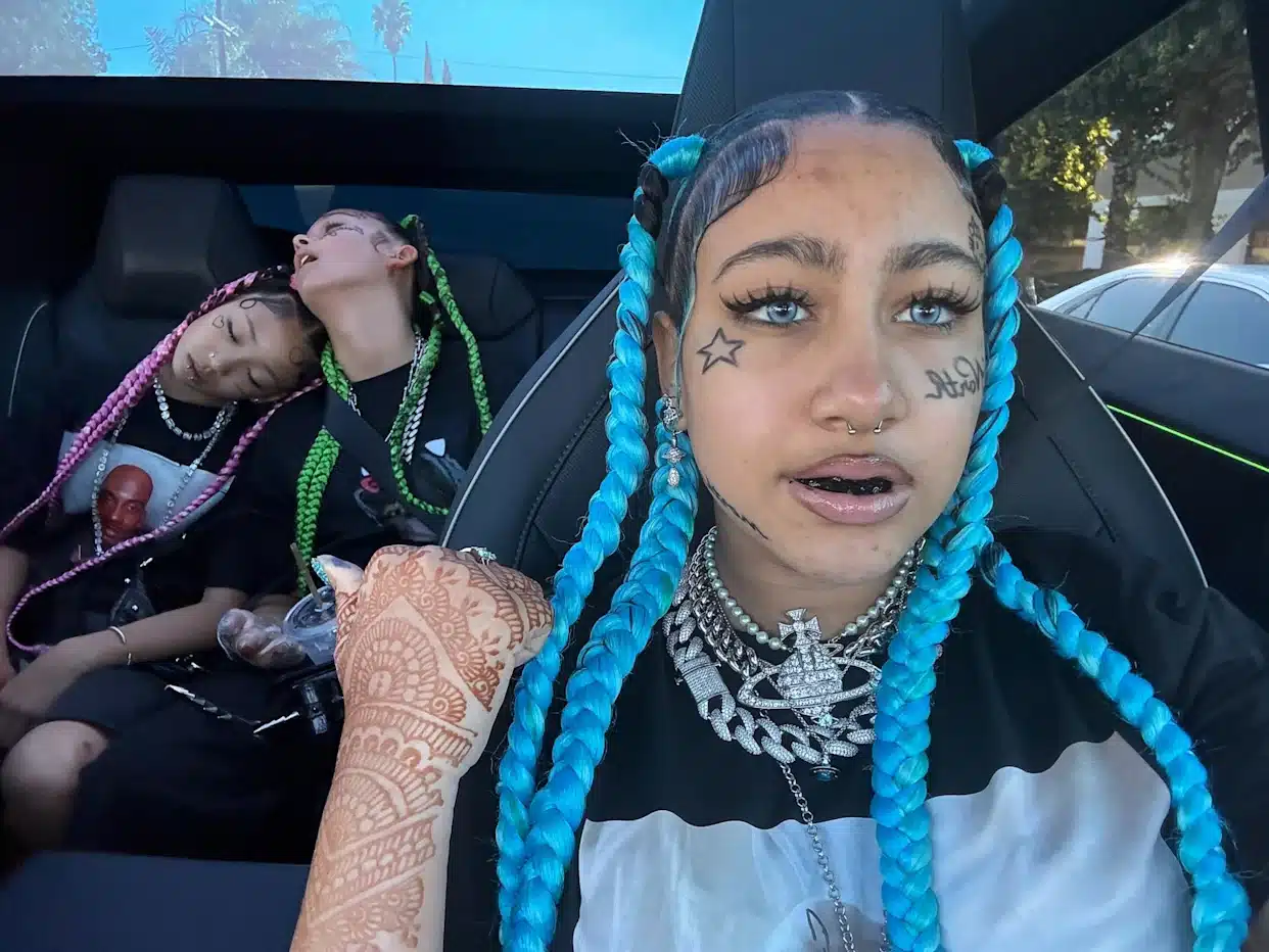 Front-seat person with bright blue braids, face tattoos, nose ring and heavy jewelry in a car; two friends sleeping in the back seat with pink and green braids.