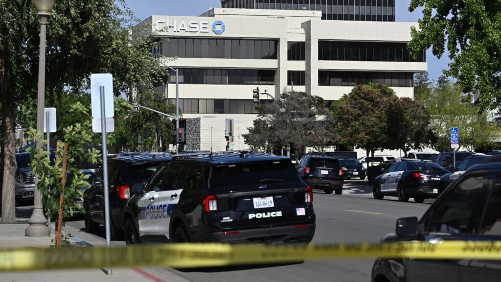 Police cars line a street in front of a Chase building, with yellow caution tape across the foreground indicating a restricted area.