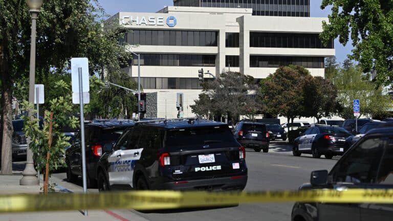 Police cars line a street in front of a Chase building, with yellow caution tape across the foreground indicating a restricted area.