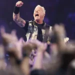 Male rock musician raises a clenched fist on stage as the crowd cheers and phones glow in the foreground.