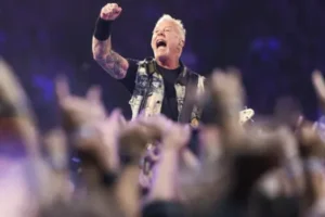 Male rock musician raises a clenched fist on stage as the crowd cheers and phones glow in the foreground.