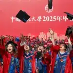 Graduates in red gowns throw their caps in the air during a University graduation ceremony against a red backdrop with Chinese text.