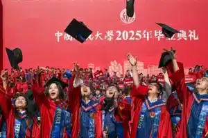 Graduates in red gowns throw their caps in the air during a University graduation ceremony against a red backdrop with Chinese text.