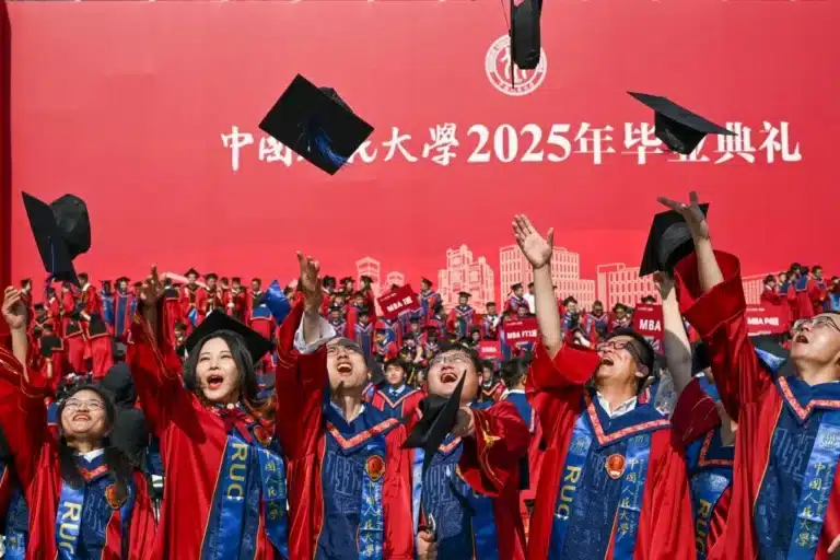 Graduates in red gowns throw their caps in the air during a University graduation ceremony against a red backdrop with Chinese text.