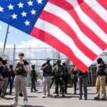 Armed security personnel in tactical gear stand near a chain-link fence with barbed wire, while a large American flag dominates the foreground.