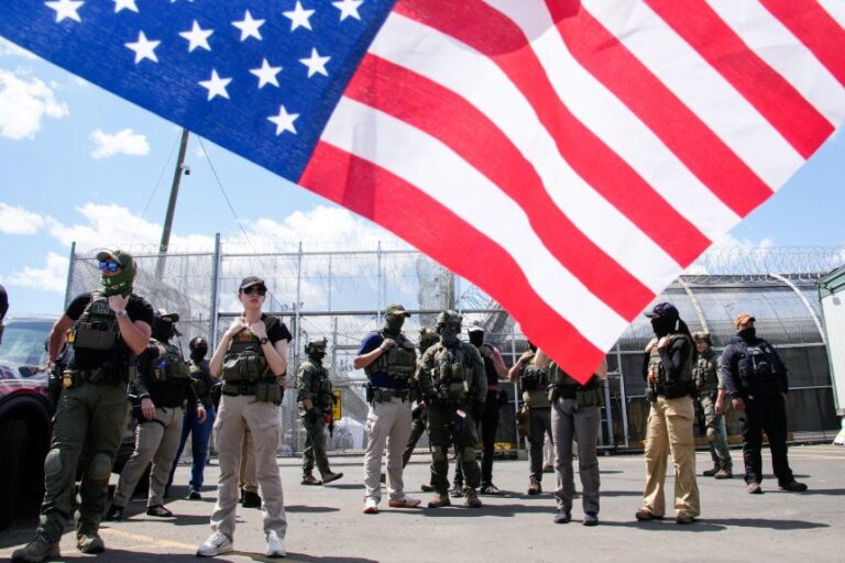 Armed security personnel in tactical gear stand near a chain-link fence with barbed wire, while a large American flag dominates the foreground.