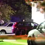 Nighttime crime scene with yellow tape, a group of people in suits by a white SUV and a dark SUV, flashing lights in the background