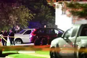 Nighttime crime scene with yellow tape, a group of people in suits by a white SUV and a dark SUV, flashing lights in the background