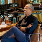 Man in a black shirt laughs into a studio microphone during a recording session at a wooden table with drinks and papers nearby, casual setting.