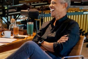 Man in a black shirt laughs into a studio microphone during a recording session at a wooden table with drinks and papers nearby, casual setting.