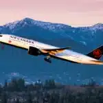 Air Canada passenger jet lifting off at sunset with snow-capped mountains and a forest below, red maple leaf on tail visible.