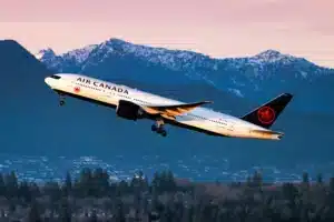 Air Canada passenger jet lifting off at sunset with snow-capped mountains and a forest below, red maple leaf on tail visible.