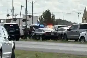 Emergency scene on a highway with police cars and flashing lights amid several parked vehicles, three or more officers assisting near the center.
