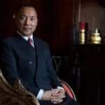 Professional portrait of a man in a navy suit seated in an ornate gold-accented chair against a dark wood-paneled backdrop.