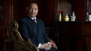 Professional portrait of a man in a navy suit seated in an ornate gold-accented chair against a dark wood-paneled backdrop.