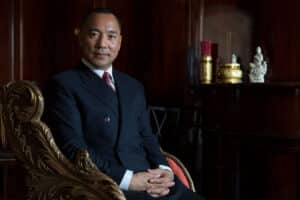 Professional portrait of a man in a navy suit seated in an ornate gold-accented chair against a dark wood-paneled backdrop.