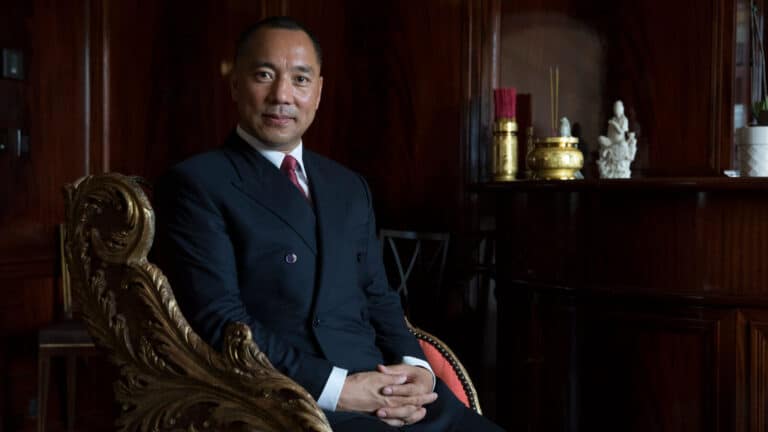 Professional portrait of a man in a navy suit seated in an ornate gold-accented chair against a dark wood-paneled backdrop.
