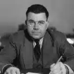Man in a suit and round glasses sits at a desk, writing with a pencil, looking toward the camera.