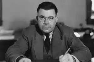 Man in a suit and round glasses sits at a desk, writing with a pencil, looking toward the camera.