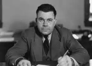 Man in a suit and round glasses sits at a desk, writing with a pencil, looking toward the camera.