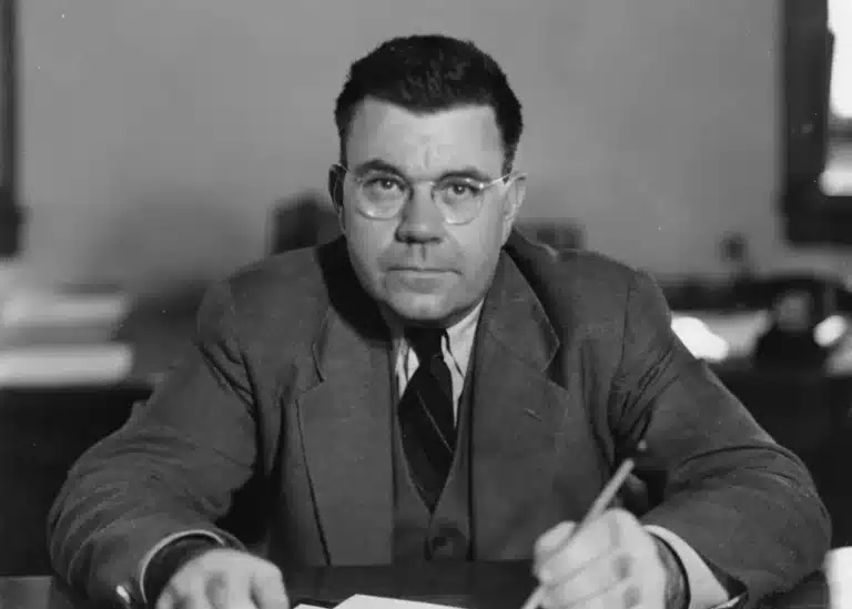 Man in a suit and round glasses sits at a desk, writing with a pencil, looking toward the camera.