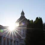 Sunlight bursts through the dome of a neoclassical government building with tall columns, flanked by trees.