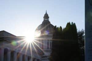 Sunlight bursts through the dome of a neoclassical government building with tall columns, flanked by trees.