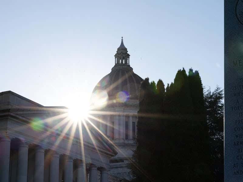 Sunlight bursts through the dome of a neoclassical government building with tall columns, flanked by trees.