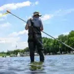 Person wearing waders stands in a shallow river holding a long pole, with the Washington Monument in the distance on a sunny day.