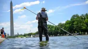 Person wearing waders stands in a shallow river holding a long pole, with the Washington Monument in the distance on a sunny day.