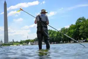 Person wearing waders stands in a shallow river holding a long pole, with the Washington Monument in the distance on a sunny day.