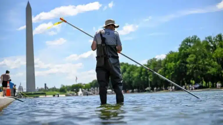 Person wearing waders stands in a shallow river holding a long pole, with the Washington Monument in the distance on a sunny day.