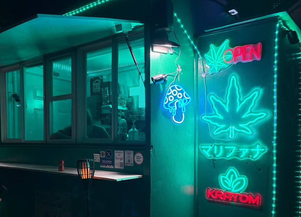 Night storefront with teal neon signs including a large leaf motif and 'KRATOM' sign, a window counter, and small lamps outside.