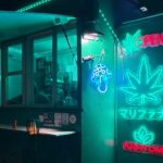 Night storefront with teal neon signs including a large leaf motif and 'KRATOM' sign, a window counter, and small lamps outside.