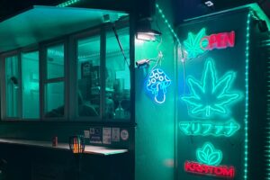 Night storefront with teal neon signs including a large leaf motif and 'KRATOM' sign, a window counter, and small lamps outside.