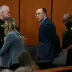 A man in a blue suit walks down a courtroom aisle flanked by two deputies in black uniforms with yellow patches.
