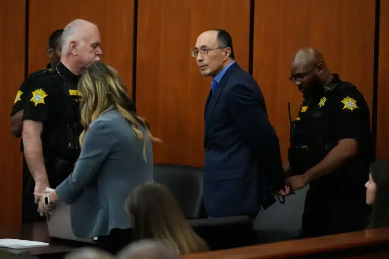A man in a blue suit walks down a courtroom aisle flanked by two deputies in black uniforms with yellow patches.