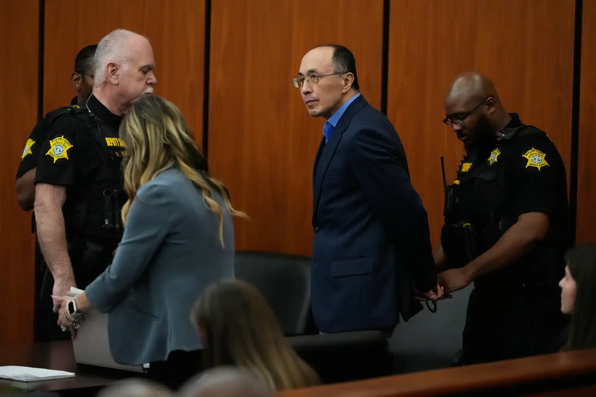A man in a blue suit walks down a courtroom aisle flanked by two deputies in black uniforms with yellow patches.