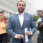 A man in a blue checkered suit walks forward, holding a folded slip of paper, as reporters photograph him in the background.