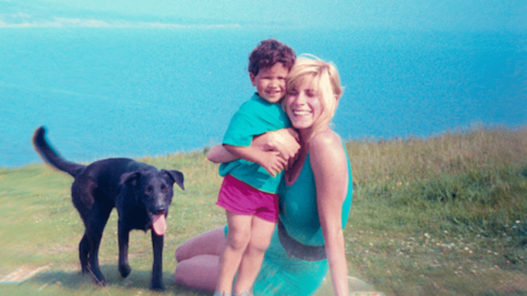 Woman holding a smiling child on a grassy hill near a black dog with a blue lake in the background.
