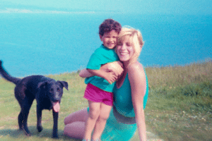 Woman holding a smiling child on a grassy hill near a black dog with a blue lake in the background.