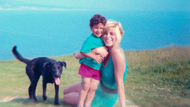 Woman holding a smiling child on a grassy hill near a black dog with a blue lake in the background.