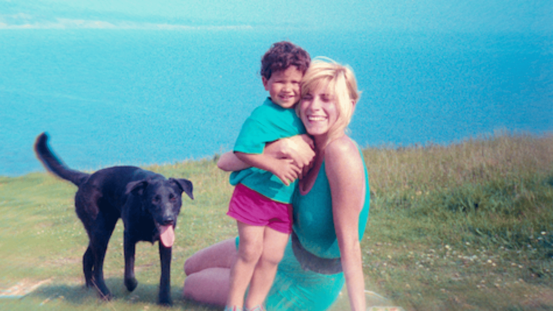 Woman holding a smiling child on a grassy hill near a black dog with a blue lake in the background.