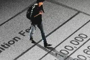 Man with a backpack walks across a surface printed with large numbers and the words resembling 'submission fee', suggesting costs or fees.