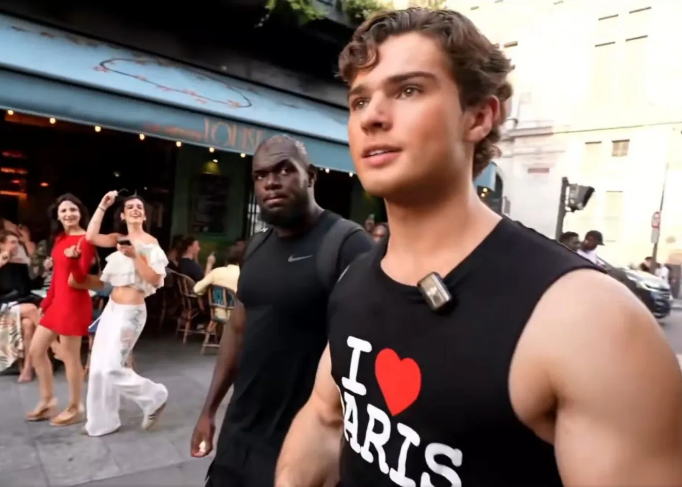 Young man in a sleeveless black shirt with an 'I ❤️ Paris' print walks on a busy street, a microphone clipped to his shirt. Behind him, people sit at an outdoor cafe and two women pose and dance.