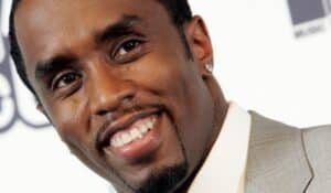 Close-up of a man in a light gray suit smiling at the camera, with a small earring visible.
