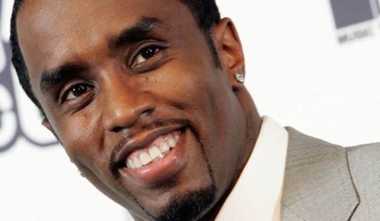 Close-up of a man in a light gray suit smiling at the camera, with a small earring visible.