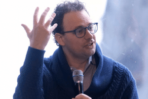Man wearing glasses and a blue knit cardigan speaks into a handheld microphone while gesturing with his left hand.